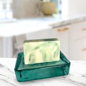 A green and cream swirled soap bar resting on a teal glass dish, placed on a marble countertop with a blurred kitchen background.