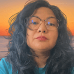 A person with long, curly black hair and glasses, looking directly at the camera, with a sunset and ocean background.